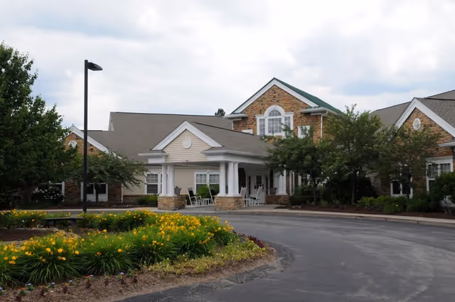 Front entrance of a senior living building with a covered porte-cochère, rocking chairs, landscaped flowerbeds, and a circular driveway.