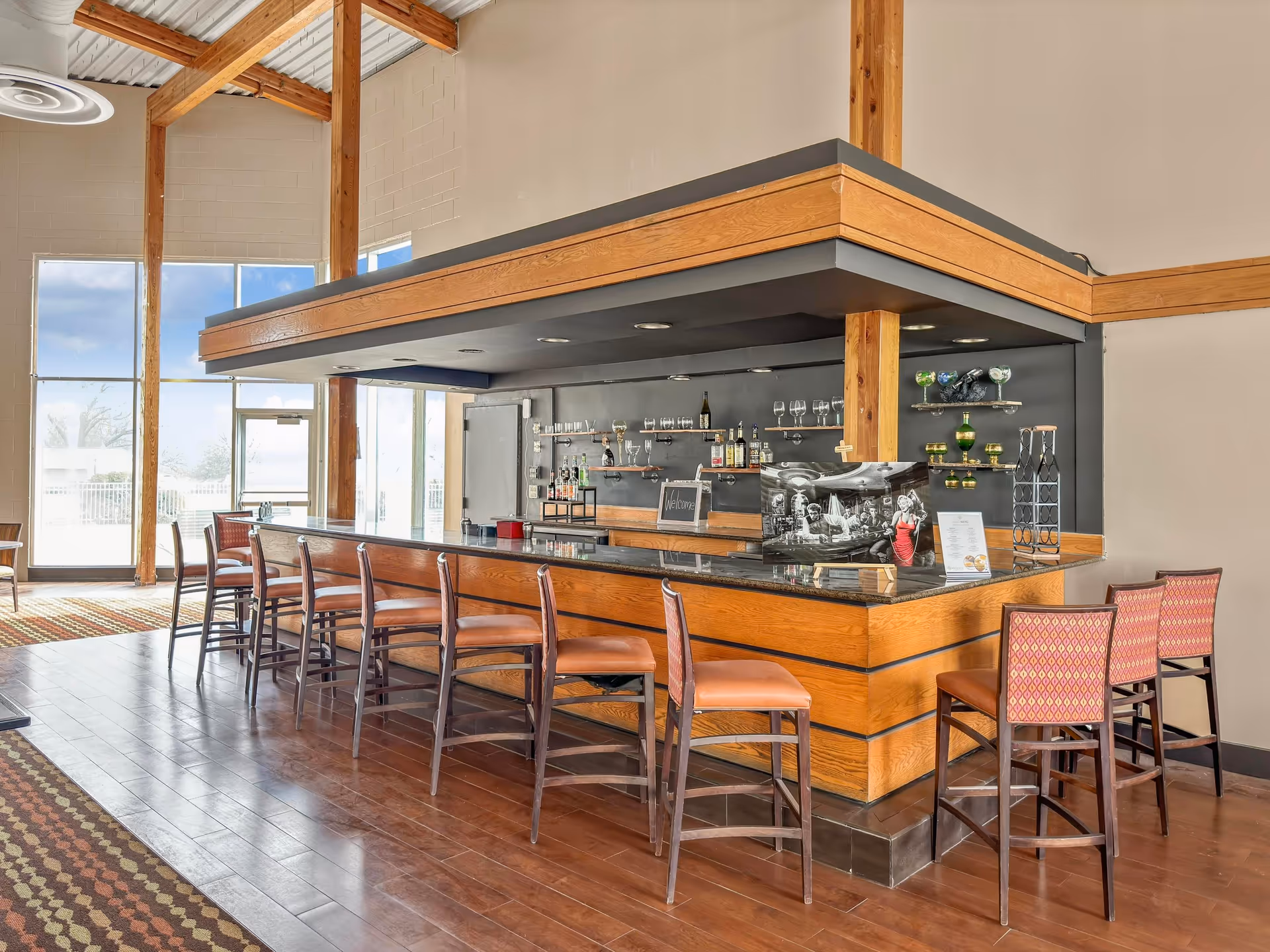 Interior bar area with a wooden counter, shelves of glassware and bottles, and several bar stools in a bright lounge space.