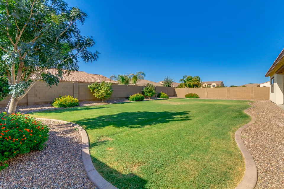 A well-maintained outdoor garden area with green grass, small bushes, and trees along a curved stone pathway, enclosed by a beige brick wall under a clear blue sky.
