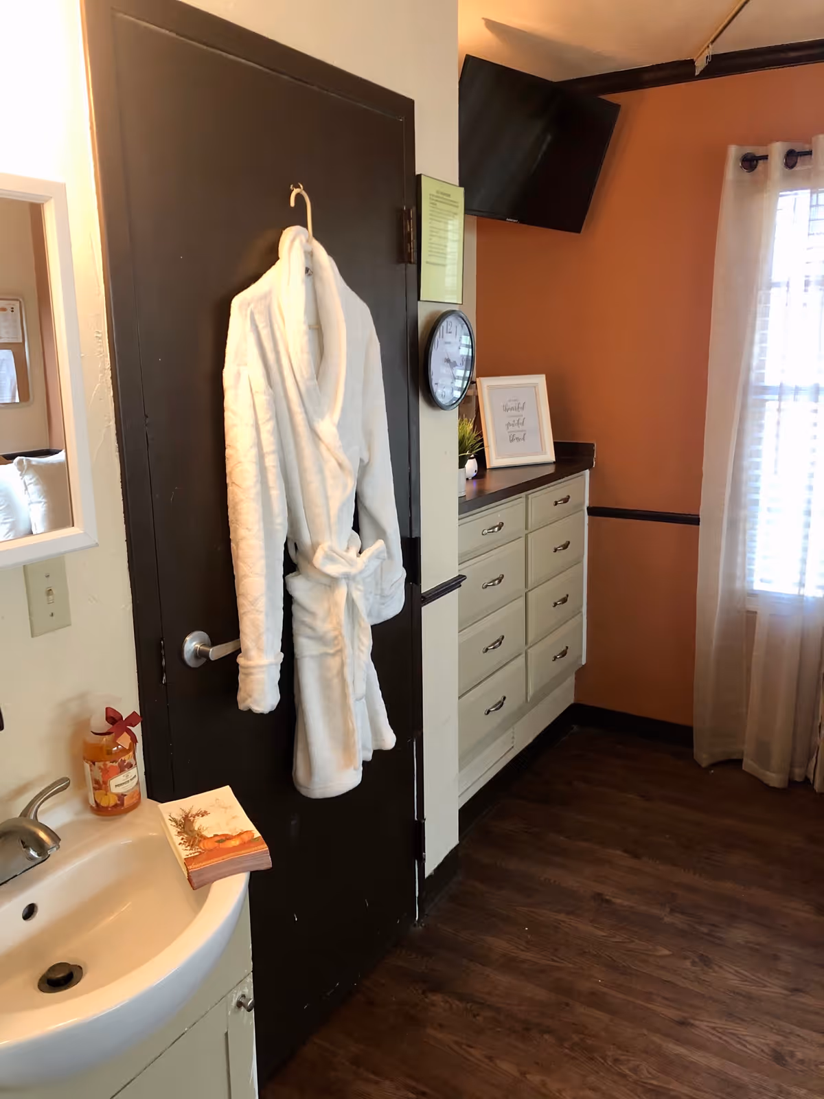 Interior view of a room with a white bathrobe hanging on a dark door. To the left, there is a small sink with a soap dispenser and a stack of napkins. On the right side, there is a set of drawers with a clock, a small plant, and a framed quote on top. A window with sheer curtains allows natural light into the room.