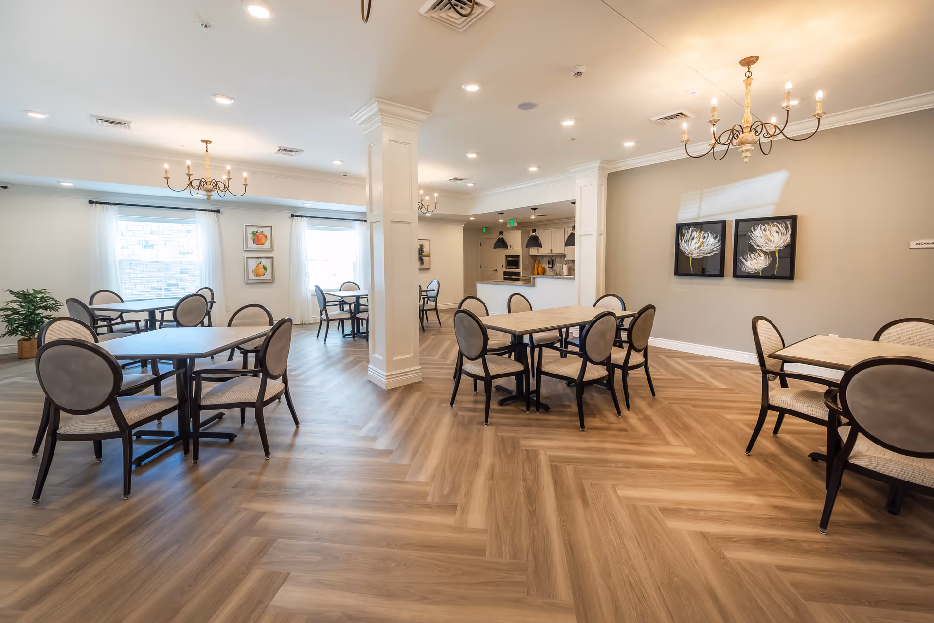 A bright and spacious dining room with multiple tables and chairs arranged neatly. The room features light-colored walls, wooden herringbone-patterned flooring, chandeliers hanging from the ceiling, and large windows with sheer white curtains allowing natural light to fill the space. There are framed floral artworks on one wall and a kitchen area visible in the background.