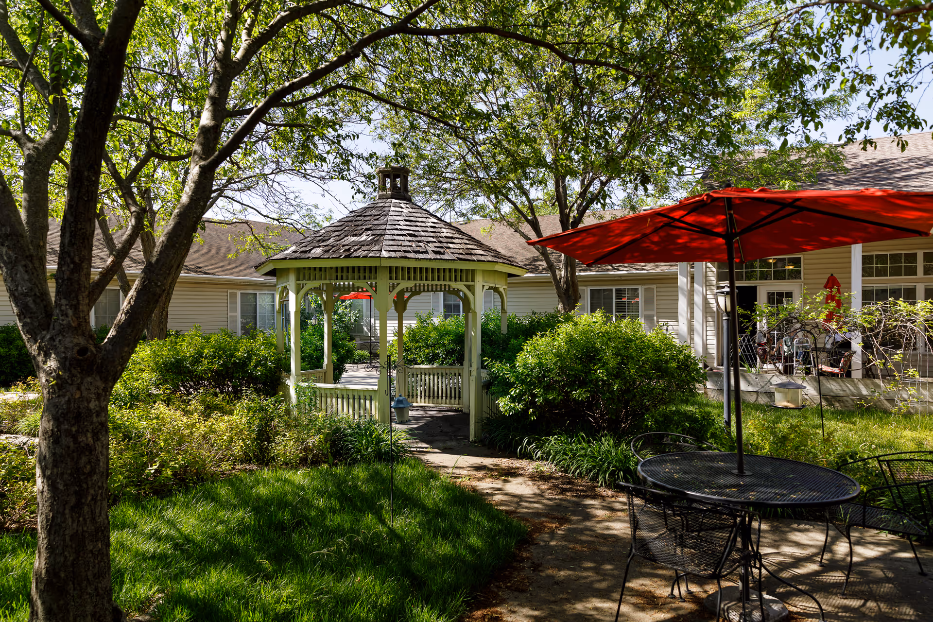 A peaceful outdoor garden area at a senior living facility featuring a white wooden gazebo surrounded by green bushes and trees. There is a black metal table with chairs and a red umbrella providing shade. The background shows a beige building with large windows and a patio area.