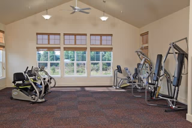 A spacious exercise room with large windows letting in natural light. The room contains various fitness equipment including stationary bikes and weight machines. The walls are light-colored and the ceiling is vaulted with ceiling fans and hanging light fixtures.