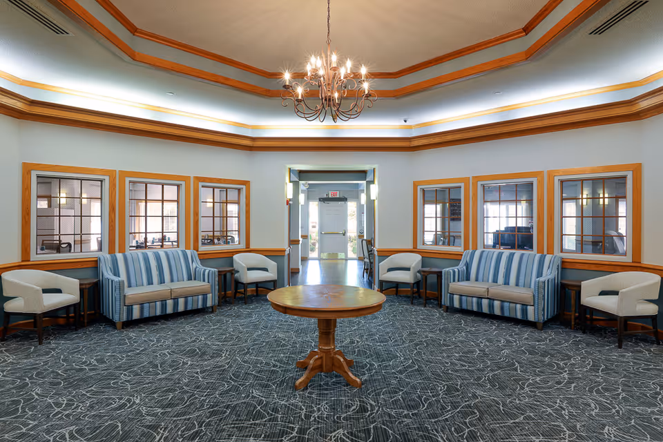 Well-lit common lounge with striped sofas and armchairs arranged around a central round wooden table under a chandelier.