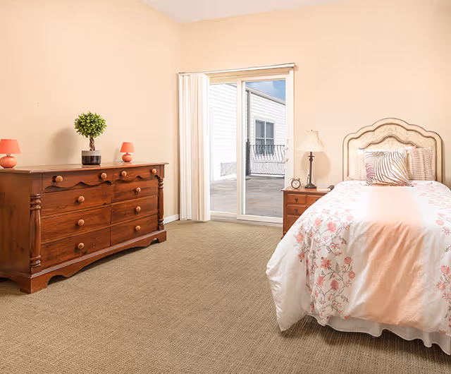 Bright bedroom with a single bed, wooden dresser, nightstand, and a sliding glass door leading outside.