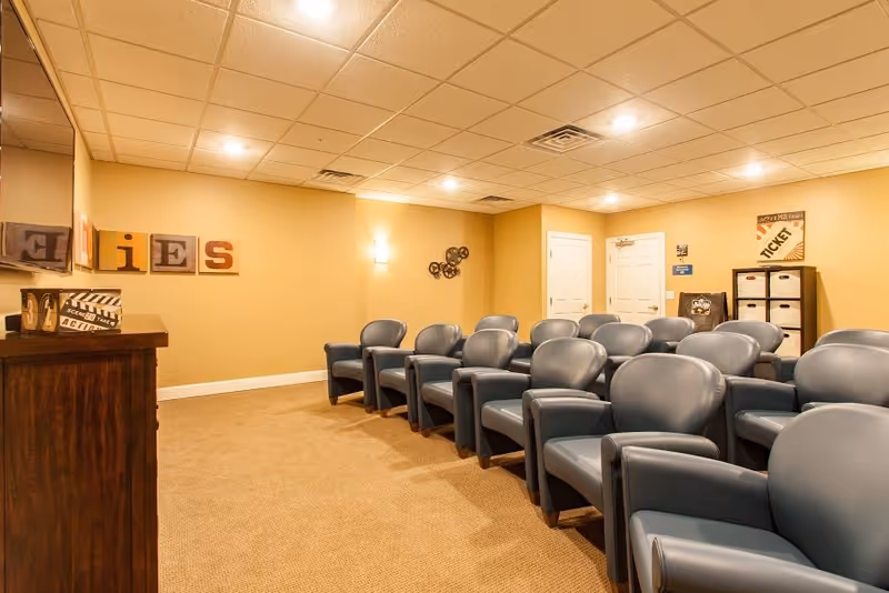 A small theater room with two rows of blue cushioned chairs facing a wall-mounted television. The walls are painted beige and decorated with movie-themed items including framed letters spelling 'TIES', a clapperboard, and a sign that says 'TICKET'. The room has a beige carpet and recessed lighting in the ceiling.