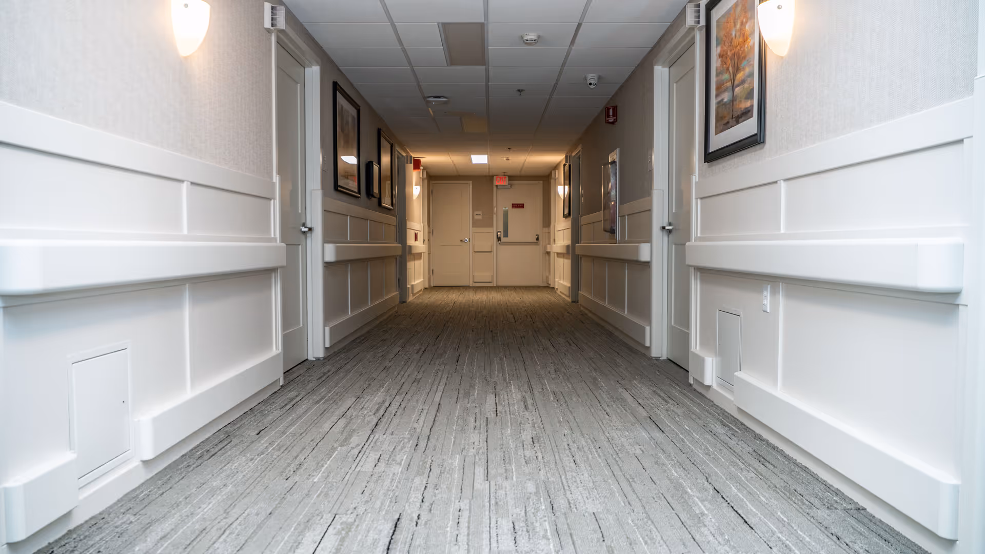 A clean, well-lit hallway in a skilled nursing and rehabilitation center with light-colored walls, carpeted floor, handrails on both sides, framed artwork on the walls, and several closed doors leading to rooms.