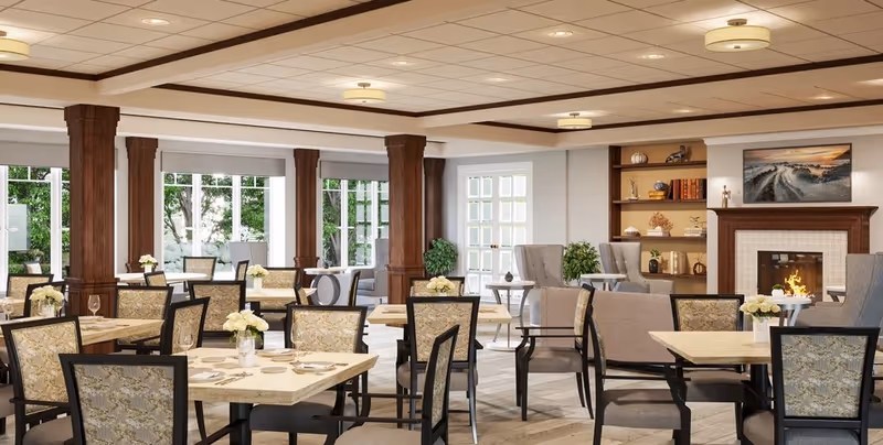 Bright senior living dining room with tables and chairs, a fireplace, bookshelves, and large windows overlooking greenery.
