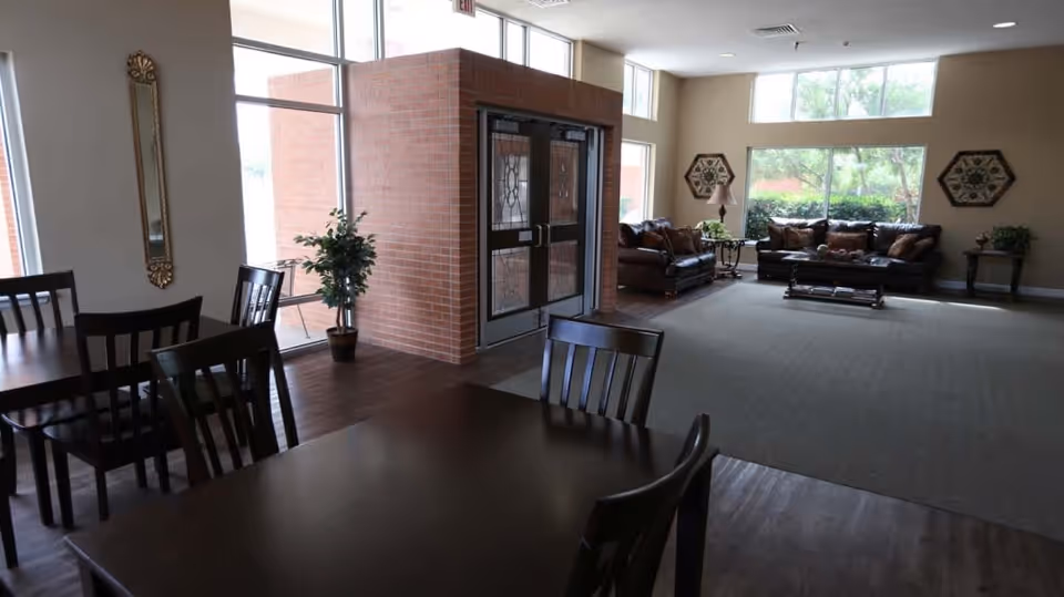 Spacious community room showing dining tables and chairs in the foreground and a lounge area with sofas by large windows in the background.
