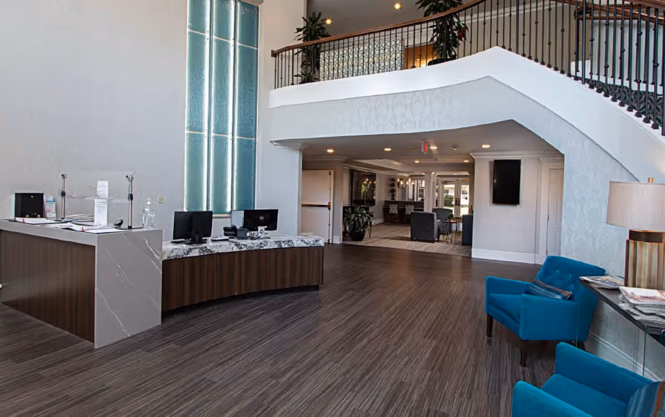 Reception area of Brooking Park facility with a curved wooden desk featuring two computer monitors, a tall decorative window behind the desk, a staircase with black railing on the right, and two blue armchairs near a side table with a lamp and magazines.