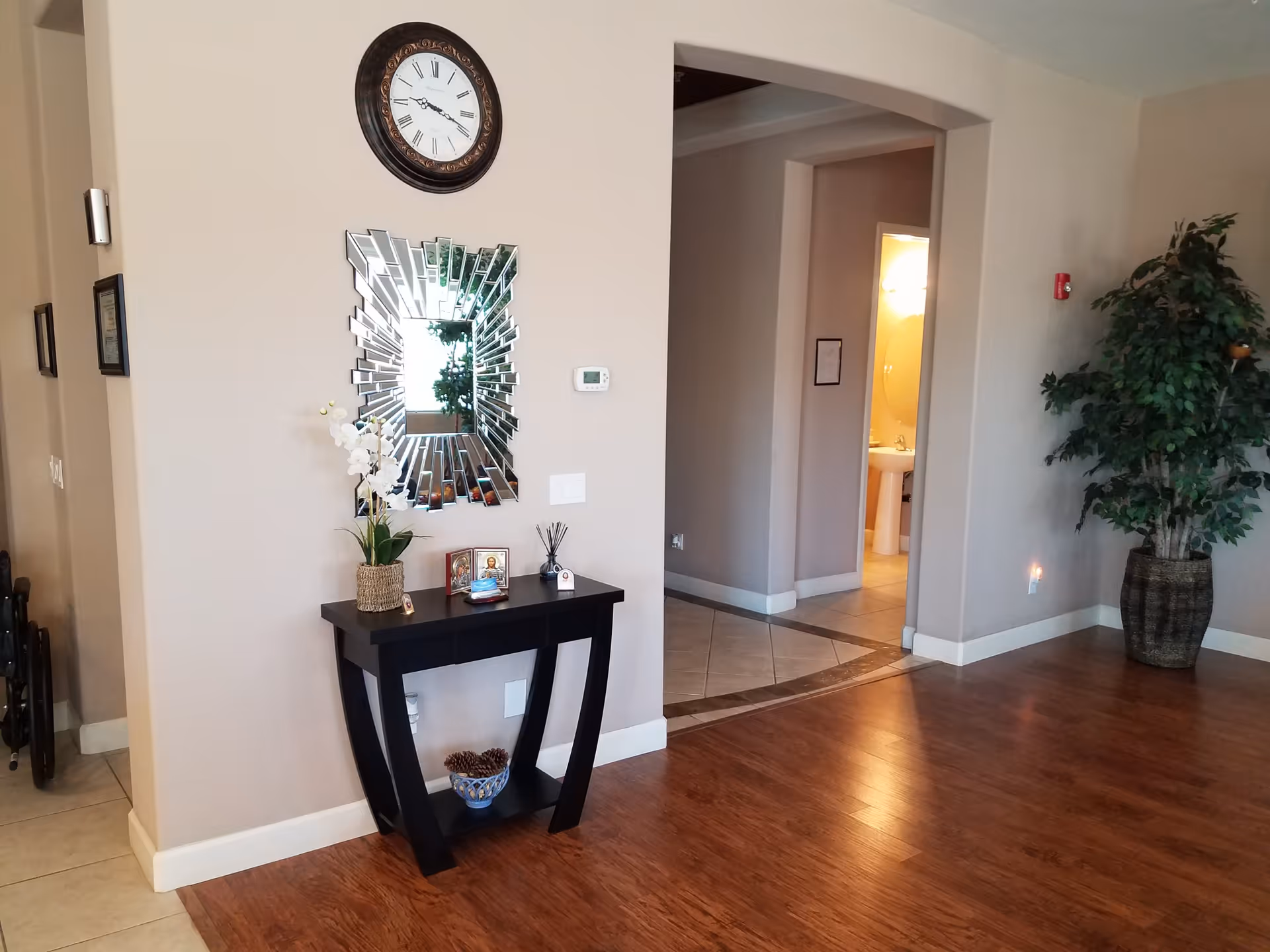 Interior view of a hallway area in a senior living facility with wooden flooring and beige walls. A decorative black console table with a potted orchid, small framed pictures, and other decor items sits against the wall beneath a large, artistic mirror and a round wall clock. To the right, there is a large potted plant near an arched doorway leading to a tiled floor area and a bathroom with a pedestal sink and mirror visible.