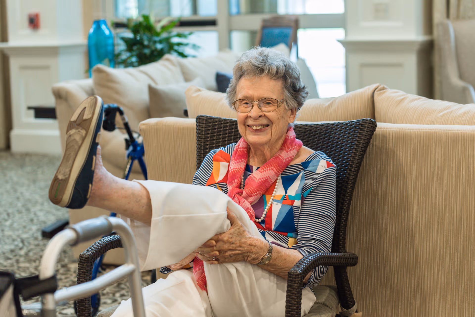 An elderly woman with glasses and a colorful top sits in a wicker chair in a cozy living room area. She is smiling and lifting one leg, showing her shoe. A walker is visible in the foreground, and a beige couch with cushions is in the background along with a blue vase and some plants.