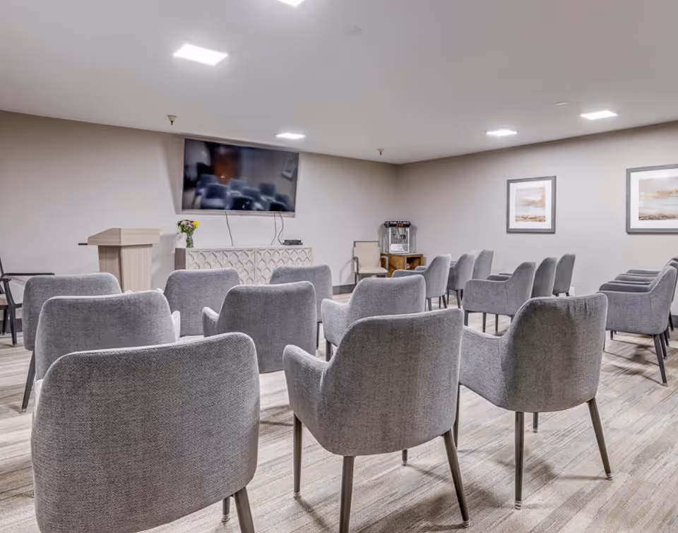 Rows of gray upholstered chairs arranged facing a wall-mounted TV, podium, and table in a meeting/activity room.