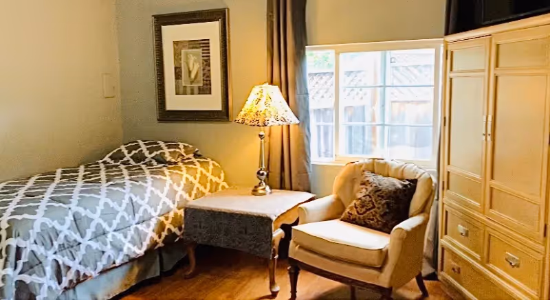 A cozy bedroom with a single bed featuring patterned bedding, a lamp-topped side table, an upholstered armchair by a window, framed art, and a large armoire.