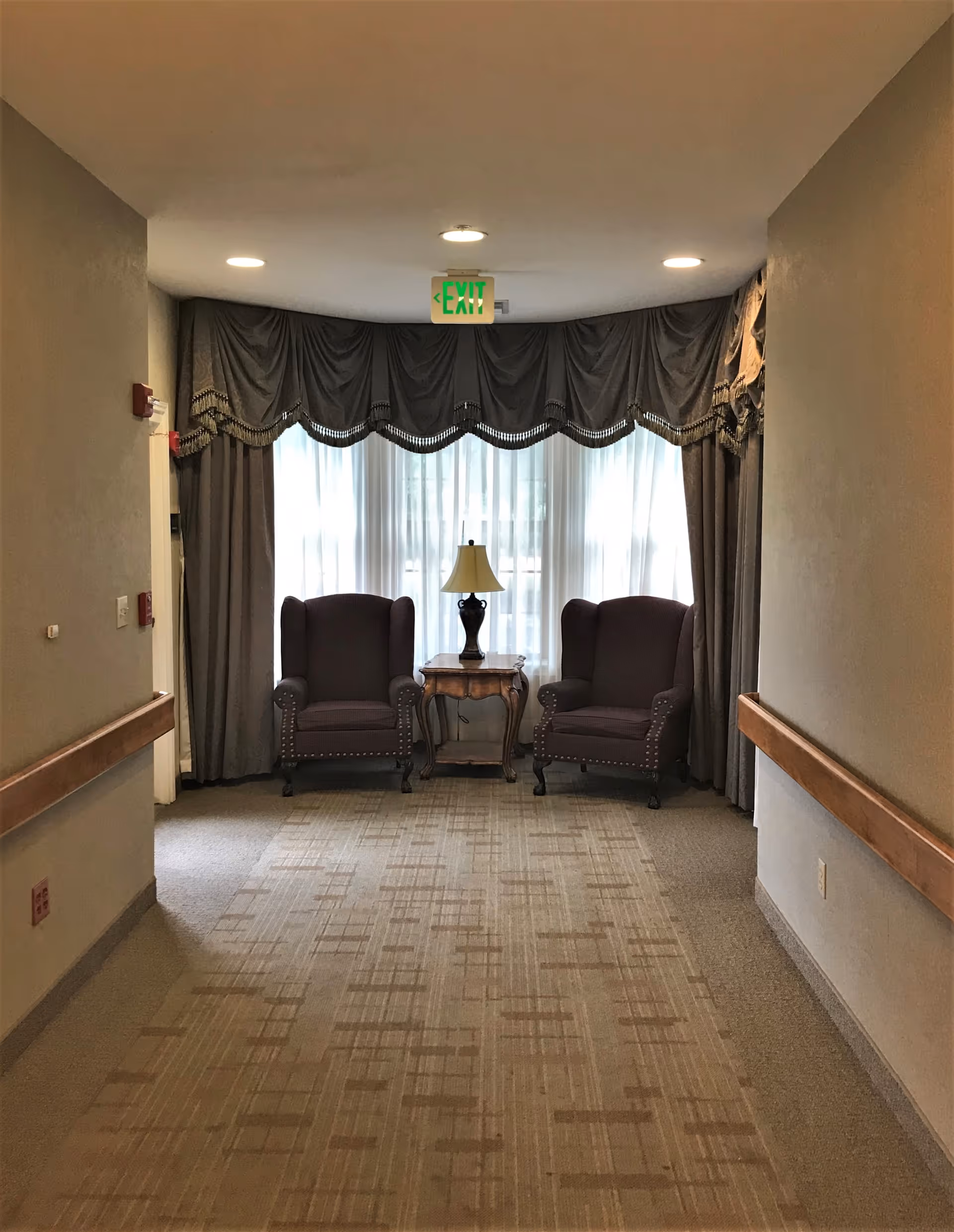 A hallway with two dark upholstered armchairs and a wooden side table with a lamp between them, positioned in front of a large window with sheer white curtains and decorative drapes. The hallway has beige walls, carpeted floor, and handrails on both sides. An illuminated green exit sign is visible above the window.