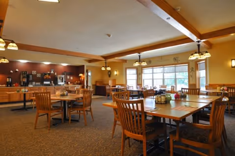 A spacious dining room with wooden tables and chairs arranged neatly. The room is well-lit with multiple hanging light fixtures and large windows allowing natural light to enter. There is a buffet area along the back wall with coffee machines and other dining amenities.