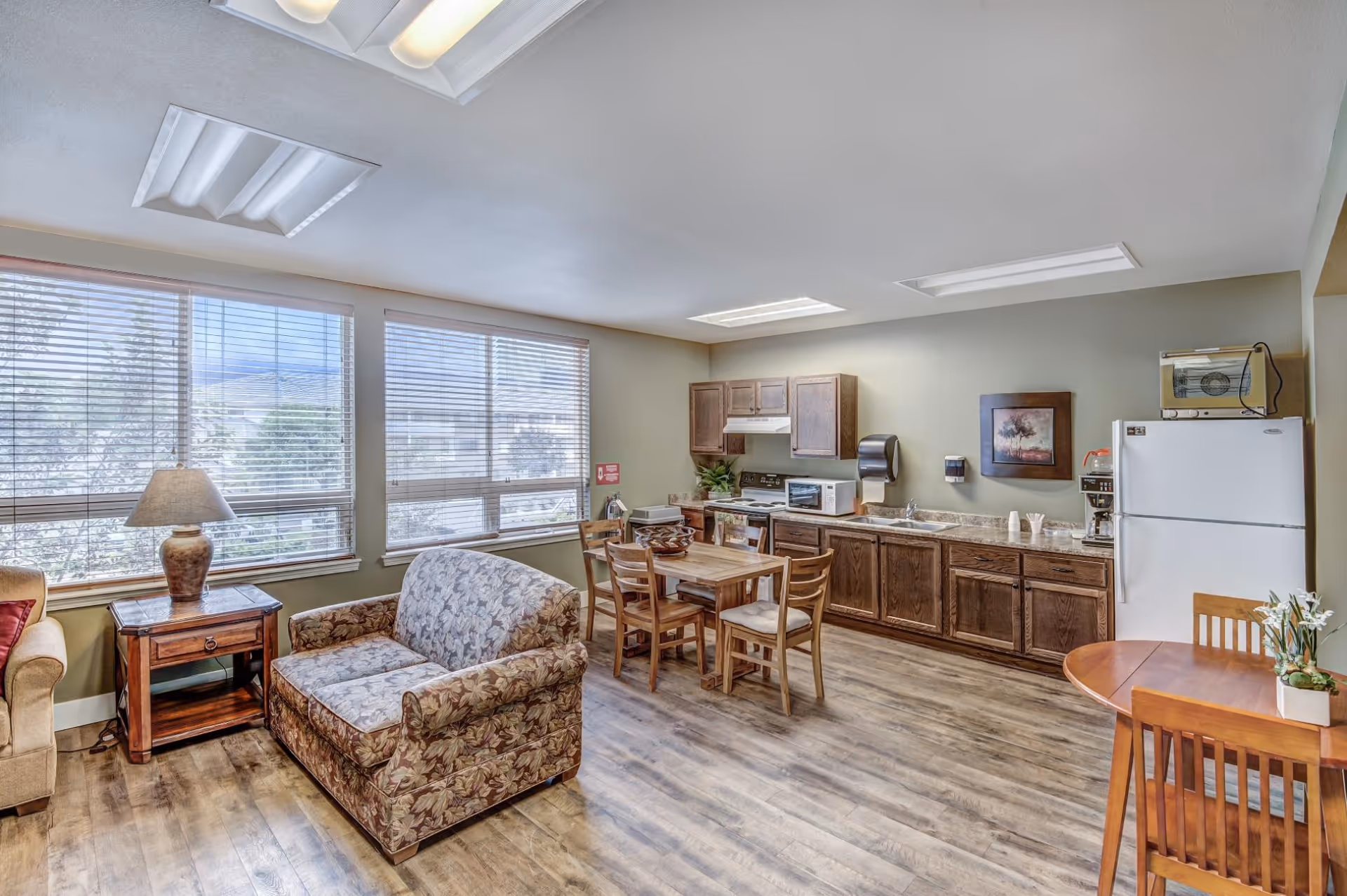 Open common living area with couches, a dining table and a small kitchenette under large windows.