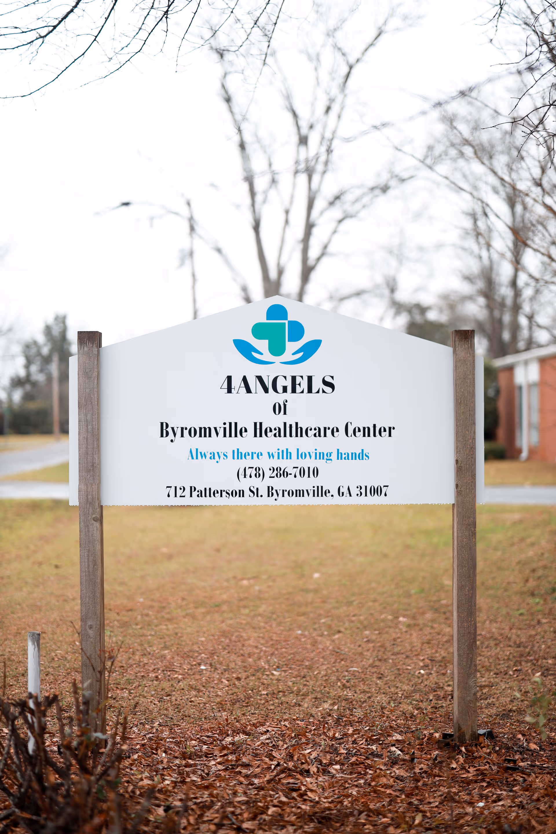 A white sign for 4ANGELS of Byromville Healthcare Center with a blue and green logo above the text. The sign includes the tagline 'Always there with loving hands' and contact information including a phone number and address. The sign is outdoors with bare trees and a grassy area with fallen leaves in the background.