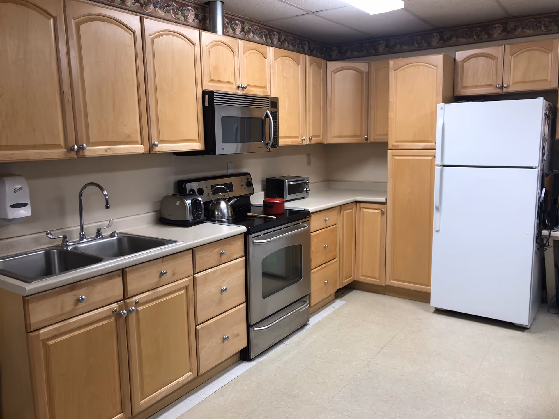 A kitchen with light wood cabinets, a double sink, a stainless steel stove with a kettle and a red pot on it, a microwave above the stove, a toaster, a small toaster oven, and a white refrigerator. The floor is tiled and there is a decorative border along the top of the wall.