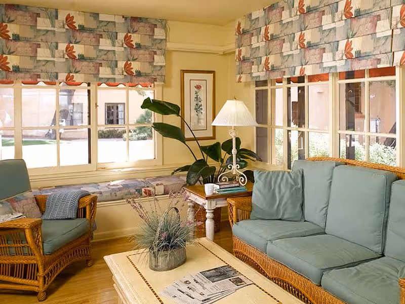 A cozy living room with wicker furniture including a sofa and armchair with green cushions, a wooden coffee table with a plant and newspapers, a side table with a lamp and books, large windows with floral valances, and a window seat with cushions.