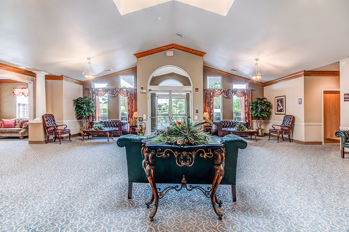 Spacious and well-lit senior living facility common area with high vaulted ceilings, patterned carpet, and elegant furniture including leather sofas, armchairs, and a decorative wooden table with a floral arrangement. Large windows with patterned curtains allow natural light to enter, and there are plants and framed artwork on the walls. Double glass doors lead outside.