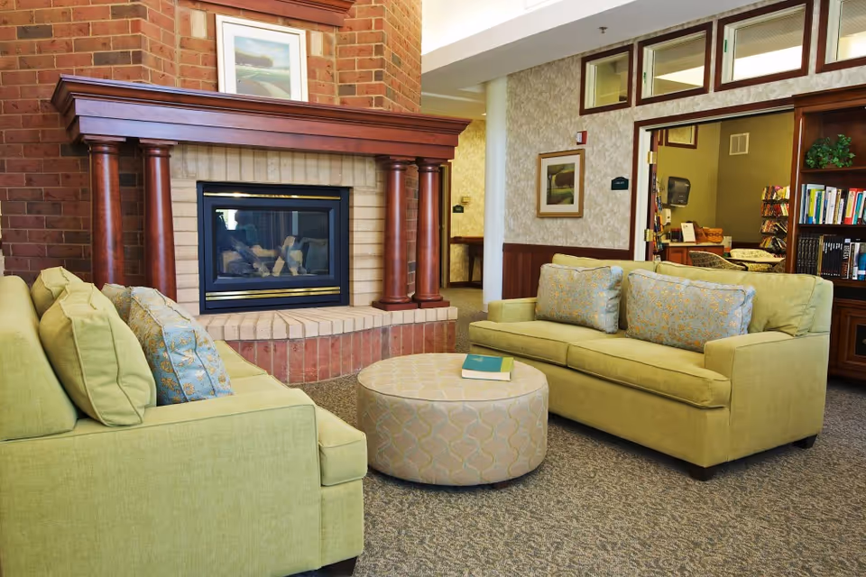 A cozy senior living facility common area with two light green sofas facing each other, a round patterned ottoman with a book on top, and a brick fireplace with wooden columns. There is a bookshelf filled with books and a framed picture on the wall.