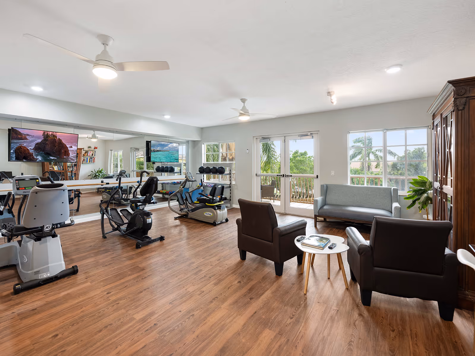 Bright multipurpose interior with exercise machines, seating area of chairs and a sofa, wall-mounted TVs and a mirrored wall, and doors opening to a balcony.