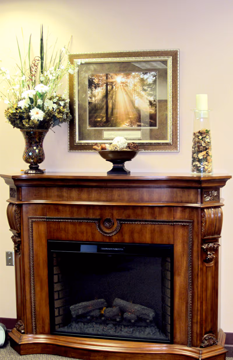 A wooden electric fireplace with ornate carvings, topped with a decorative bowl of potpourri, a tall glass vase filled with dried botanicals and a candle, and a large floral arrangement in a mosaic vase. Above the fireplace hangs a framed picture of sunlight streaming through trees in a forest.