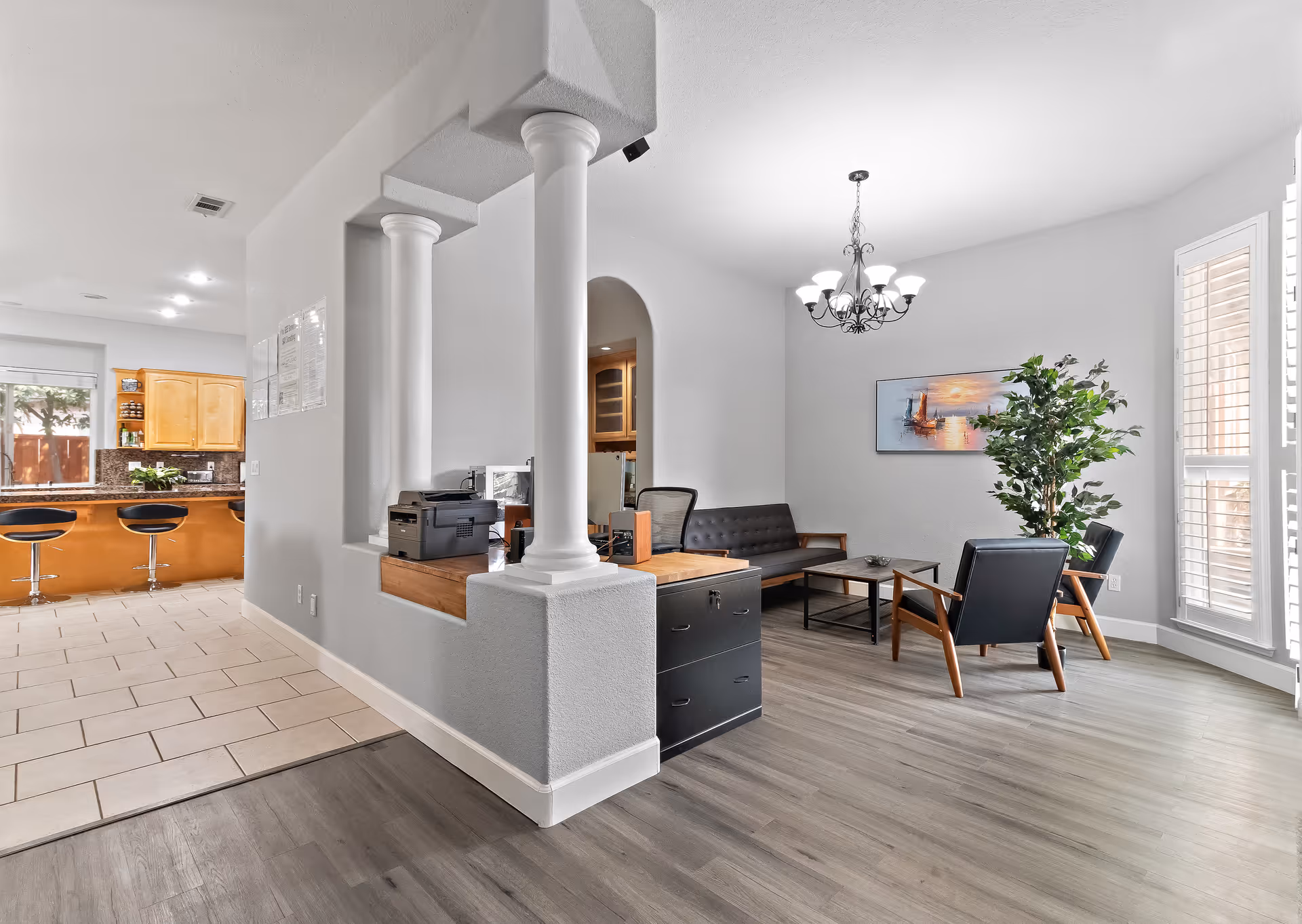 Bright and modern interior space featuring a small office area with a printer and desk, adjacent to a cozy living room with a black sofa, two black armchairs, a coffee table, a large potted plant, and a chandelier. The kitchen with wooden cabinets and bar stools is visible in the background.