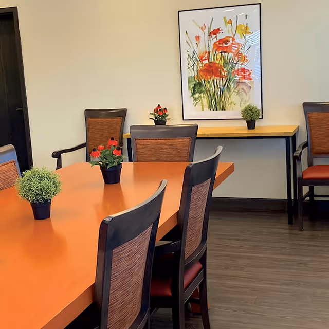 A dining room with a long wooden table surrounded by chairs with woven backs. Small potted plants with green leaves and red flowers are placed on the table and a side table against the wall. A colorful floral painting hangs on the wall above the side table. The floor is wood and the walls are light-colored.