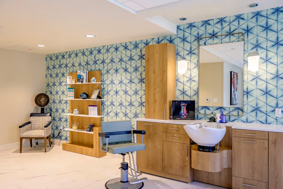 A senior living facility salon area with a hair washing station, a salon chair, a patterned blue and white wall, wooden cabinets, a large mirror, and a waiting chair next to a small shelving unit with decorative items and books.