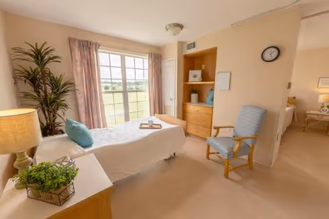 Sunlit private bedroom with a single bed, armchair, dresser, bedside table with lamp, and a large window overlooking greenery.