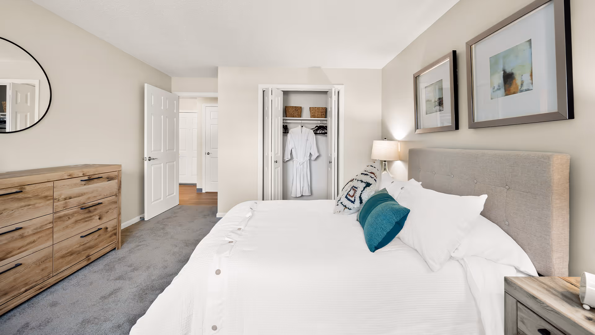 Well-lit bedroom with a neatly made bed, bedside lamp, dresser, and an open closet showing robes.