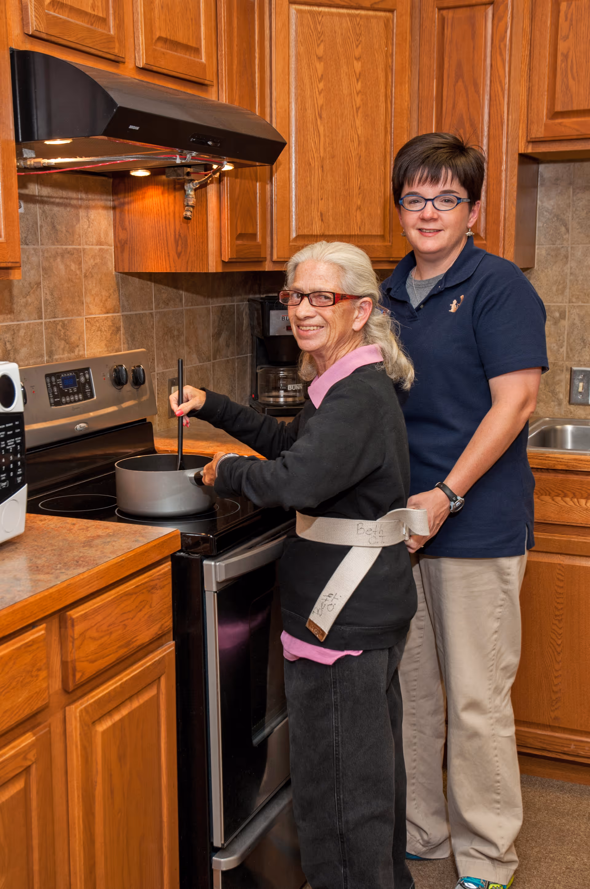 An elderly woman wearing glasses and a safety belt is stirring a pot on a stove in a kitchen, while a caregiver stands behind her providing support. The kitchen has wooden cabinets, a microwave, and a coffee maker visible in the background.
