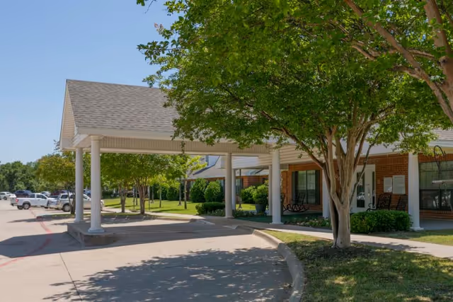 Covered entrance to a brick building with white pillars and a tree in the foreground, surrounded by green shrubs and a parking lot with several cars in the background under a clear blue sky.