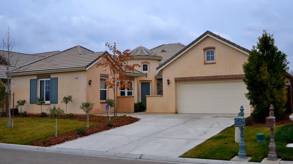 Exterior view of a single-story residential building with beige stucco walls, a tiled roof, a two-car garage, and a driveway. The front yard has small trees and shrubs planted in mulch beds, with a green lawn on either side of the driveway. The sky is overcast.