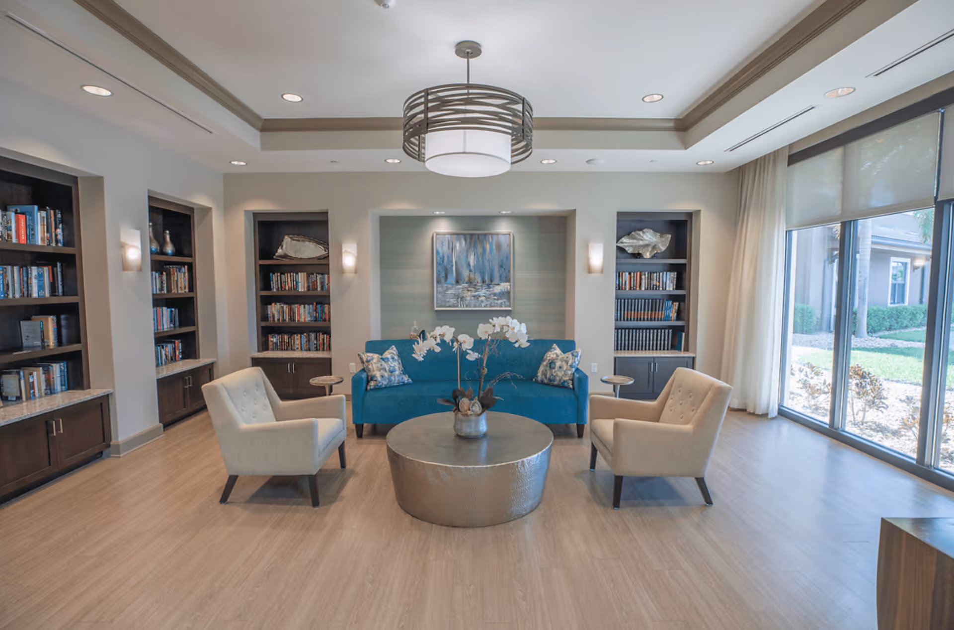 A bright and modern living room area with a blue sofa, two beige armchairs, a round metallic coffee table with a flower arrangement, built-in bookshelves filled with books and decorative items, large windows with sheer curtains letting in natural light, and a contemporary ceiling light fixture.