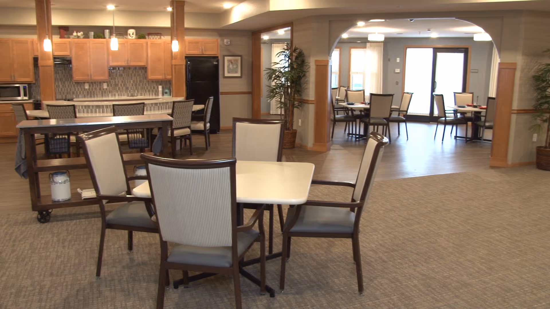A spacious dining area in a senior living facility featuring multiple tables with chairs arranged around them. In the background, there is a kitchen area with wooden cabinets, a black refrigerator, a microwave, and a tiled backsplash. The room has carpeted and hardwood flooring sections, with potted plants and soft lighting creating a warm atmosphere.