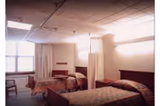 Shared resident bedroom with two single beds separated by privacy curtains, bedside tables, and a window.