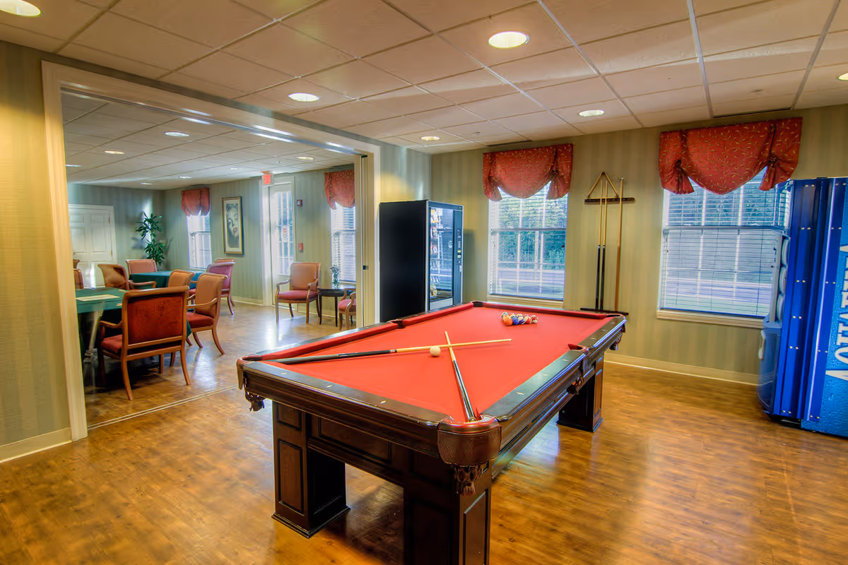 Interior view of a recreational room featuring a red felt pool table with pool cues and balls arranged on it. The room has wooden flooring, two windows with red valances, a vending machine, and a rack holding pool cues. Adjacent to this room is a seating area with several chairs and tables, decorated with framed artwork and plants.