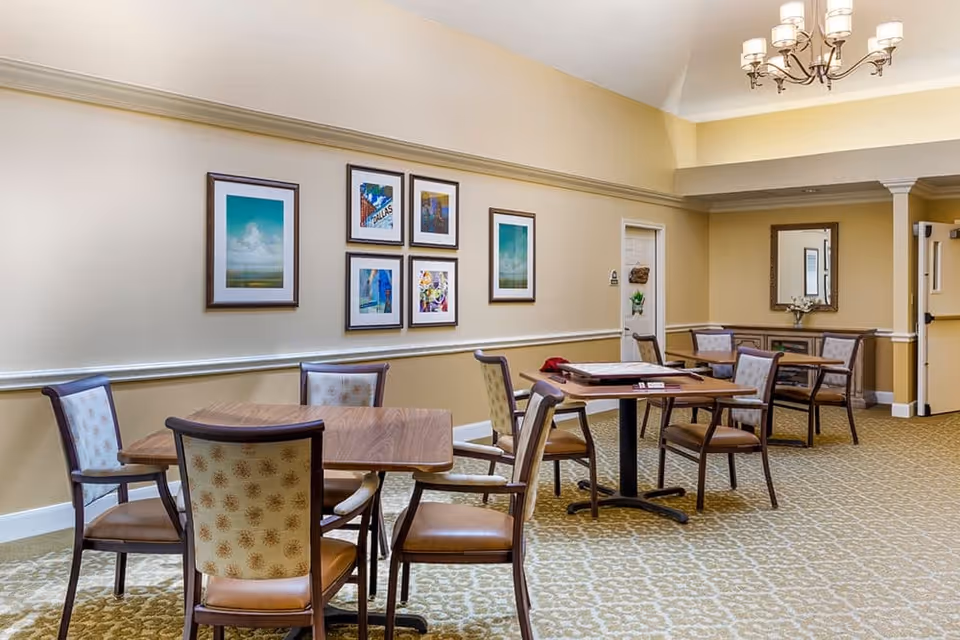 A well-lit room with several wooden tables and cushioned chairs arranged for seating. The walls are painted in a light beige color with framed artwork hanging. A chandelier with multiple lights hangs from the ceiling. There is a mirror on the far wall above a small cabinet, and a door is visible in the background.