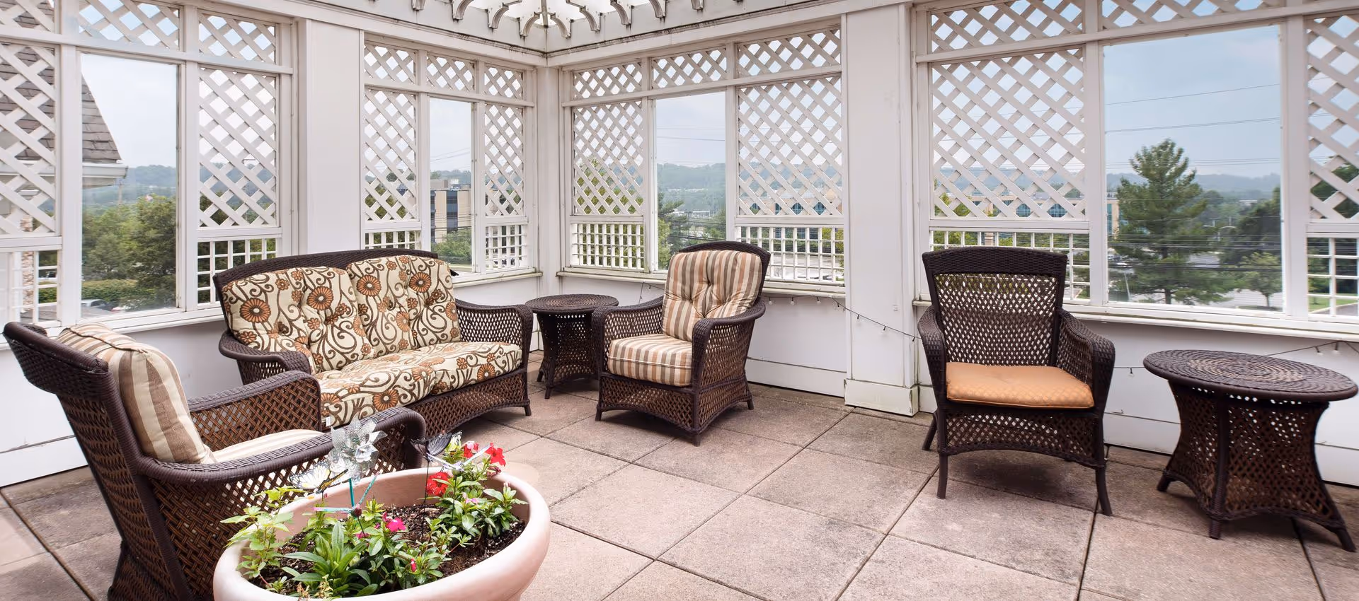 A covered outdoor patio area with wicker furniture including a cushioned loveseat, two cushioned armchairs, a wicker chair with a cushion, two small wicker tables, and a large planter with flowers. The patio has white lattice walls and a view of trees and buildings in the background.