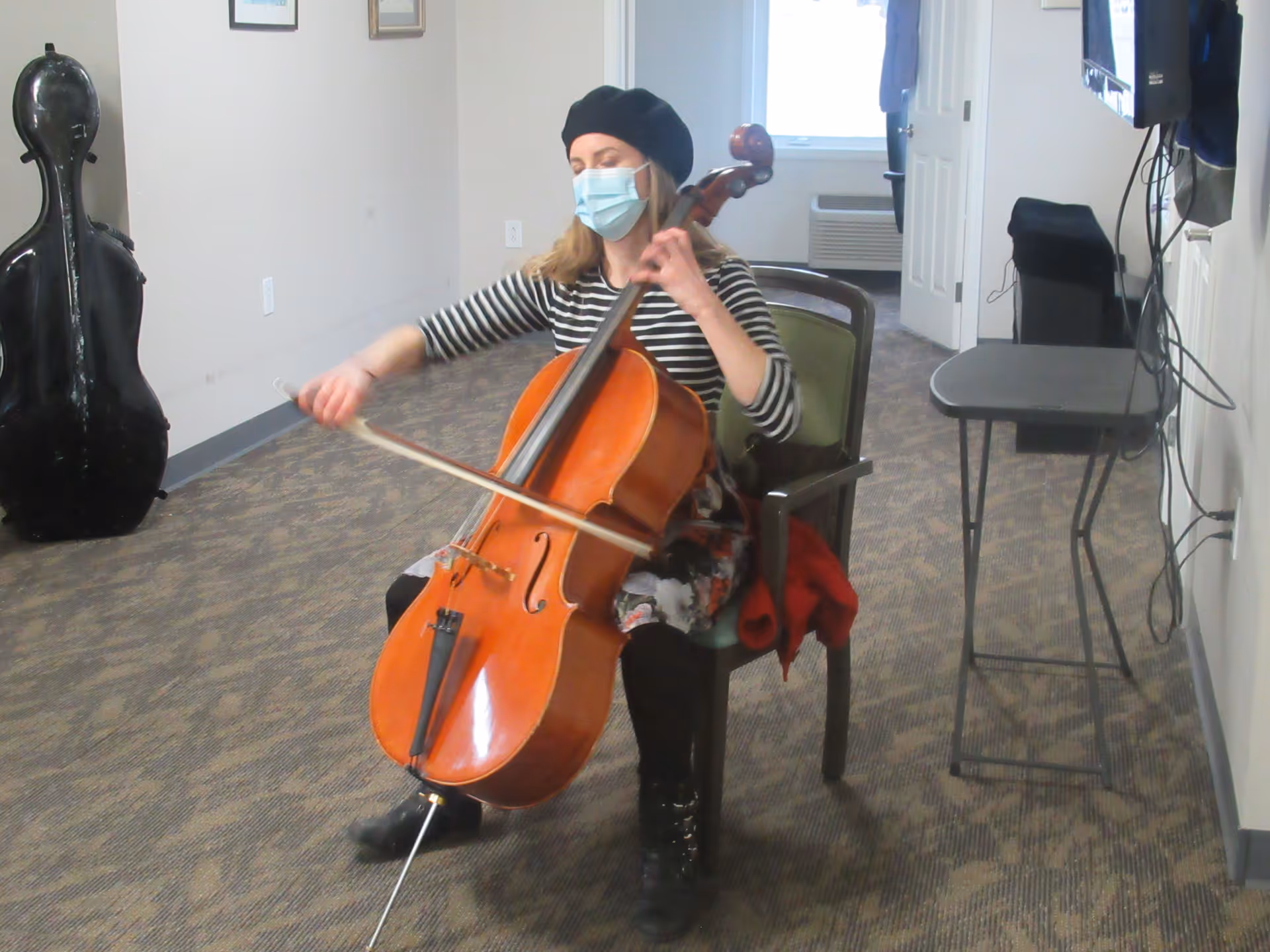 A person wearing a black beret and a face mask is seated on a chair playing a cello in a carpeted room with light-colored walls. There is a cello case standing against the wall and a small table with wires on the right side. A window and an air conditioning unit are visible in the background.