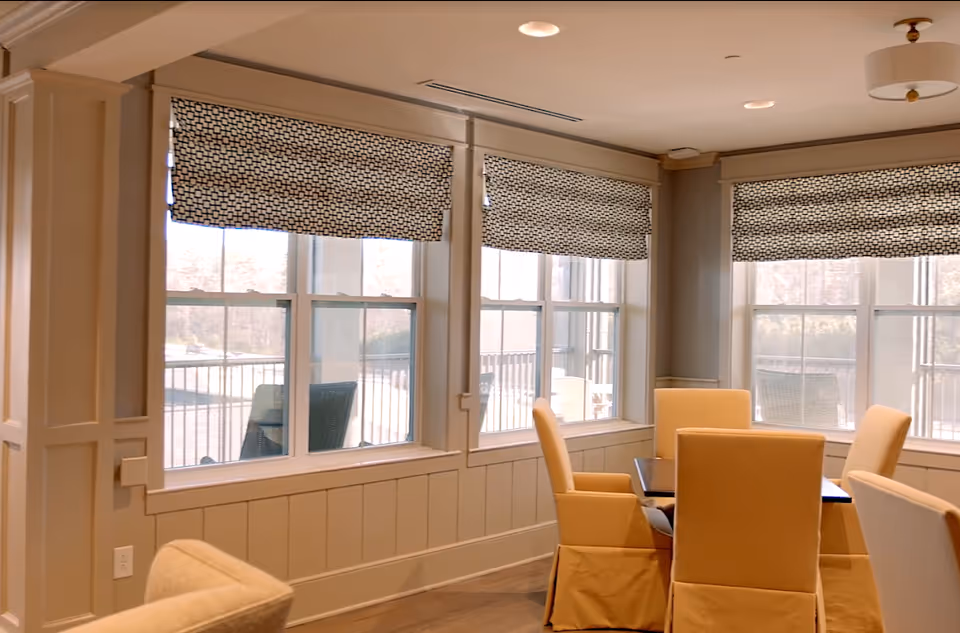 Bright dining area with a table and upholstered chairs by large windows fitted with patterned Roman shades.