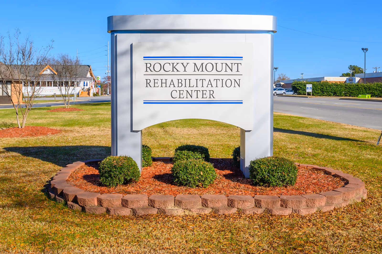 Outdoor view of a large white sign with blue accents that reads 'Rocky Mount Rehabilitation Center' placed on a landscaped area with small bushes and mulch, next to a road with buildings and cars in the background under a clear blue sky.