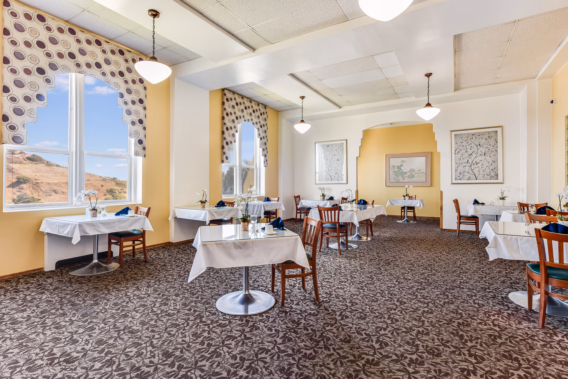 A bright dining room with several tables covered in white tablecloths and set with blue napkins and small flower arrangements. The room has large windows with patterned valances, yellow and white walls, framed artwork, and pendant lights hanging from the ceiling. The carpet has a leaf pattern.