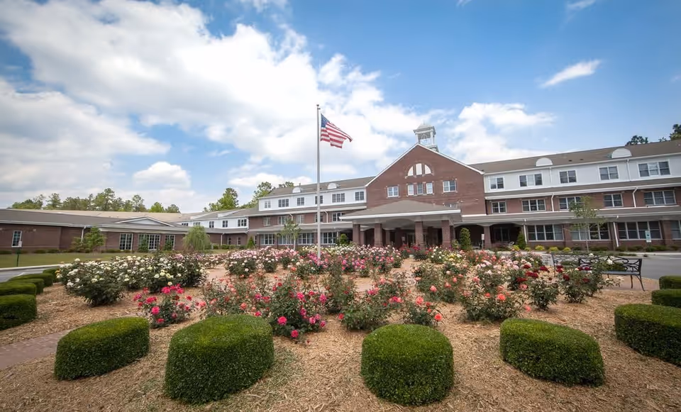 Front exterior view of a large brick residential building with a covered entrance, an American flag on a flagpole, and a landscaped garden with blooming flowers and trimmed bushes in the foreground under a partly cloudy sky.