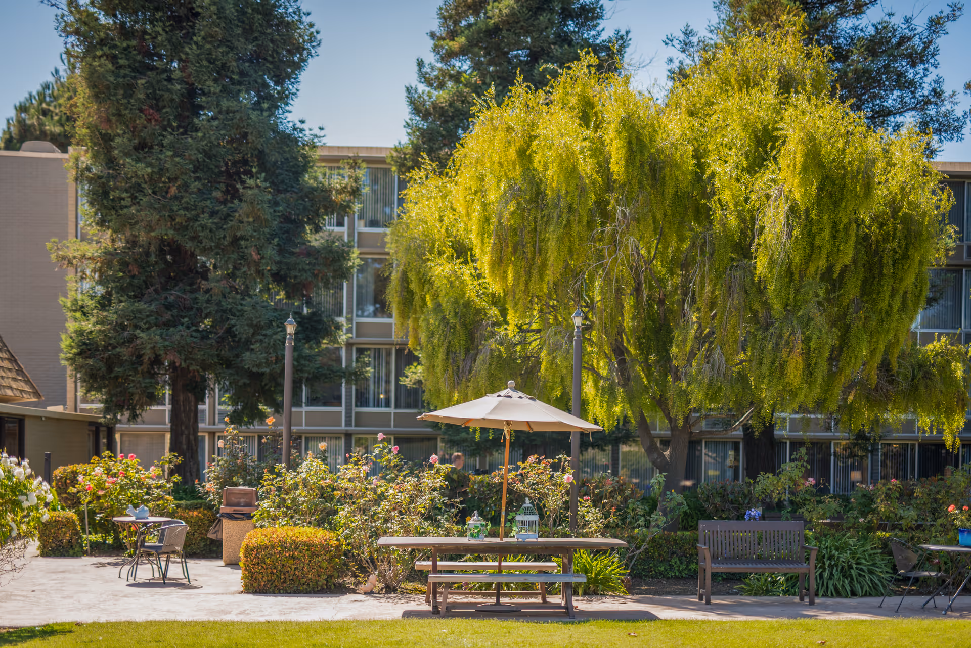 Outdoor garden area at Santa Maria Terrace featuring a picnic table with an umbrella, benches, and various plants and trees, with a multi-story building in the background.