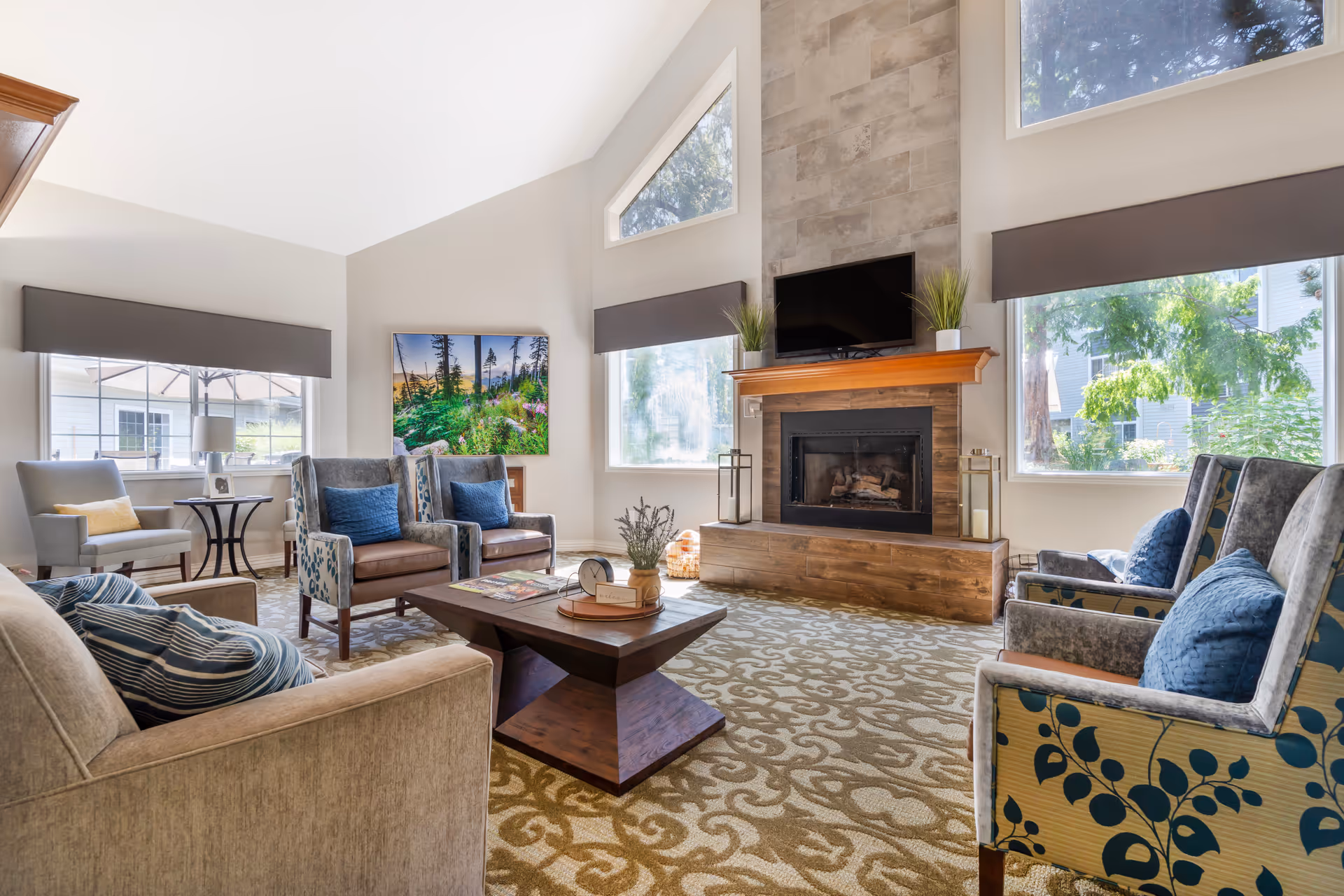 A bright and spacious living room with large windows allowing natural light to fill the space. The room features a fireplace with a mounted TV above it, two potted plants on the mantel, and two lanterns on either side of the fireplace. There are multiple upholstered chairs with blue cushions, a beige sofa with striped pillows, a wooden coffee table with decorative items, and a side table with a lamp. The carpet has a patterned design, and a colorful nature-themed painting hangs on the wall.