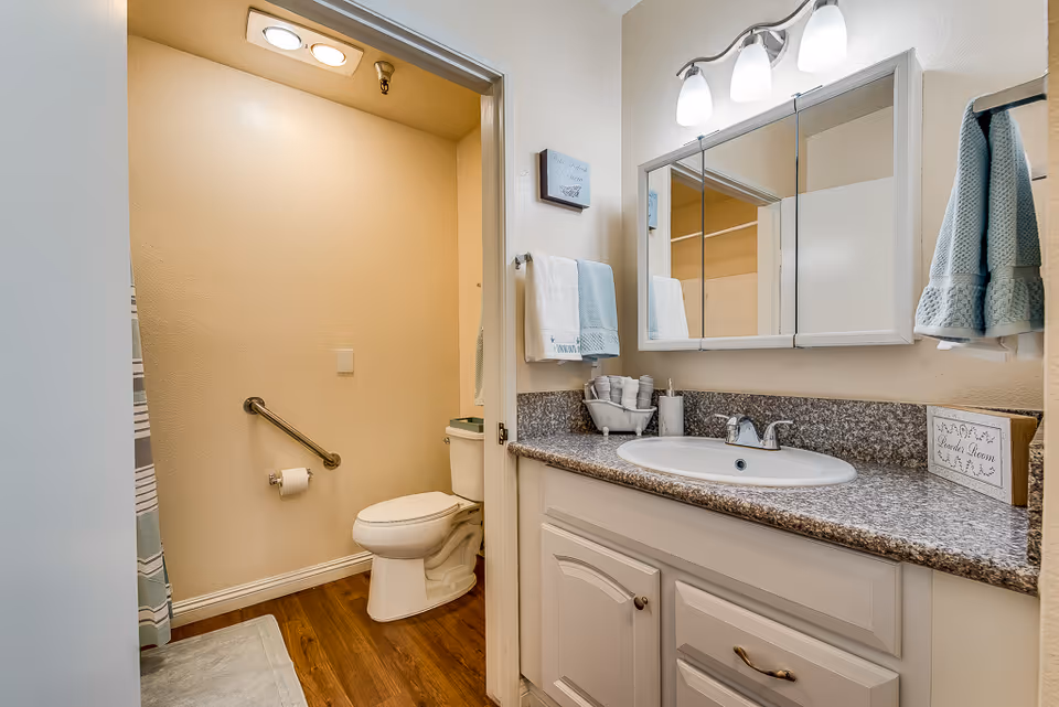 A clean and well-lit bathroom with a white toilet, a grab bar on the wall, and a toilet paper holder. The bathroom has wood flooring and beige walls. To the right, there is a vanity with a granite countertop, a white sink, a large mirror with three lights above it, and neatly folded towels hanging on the wall. A small decorative sign labeled 'Powder Room' is placed on the countertop.